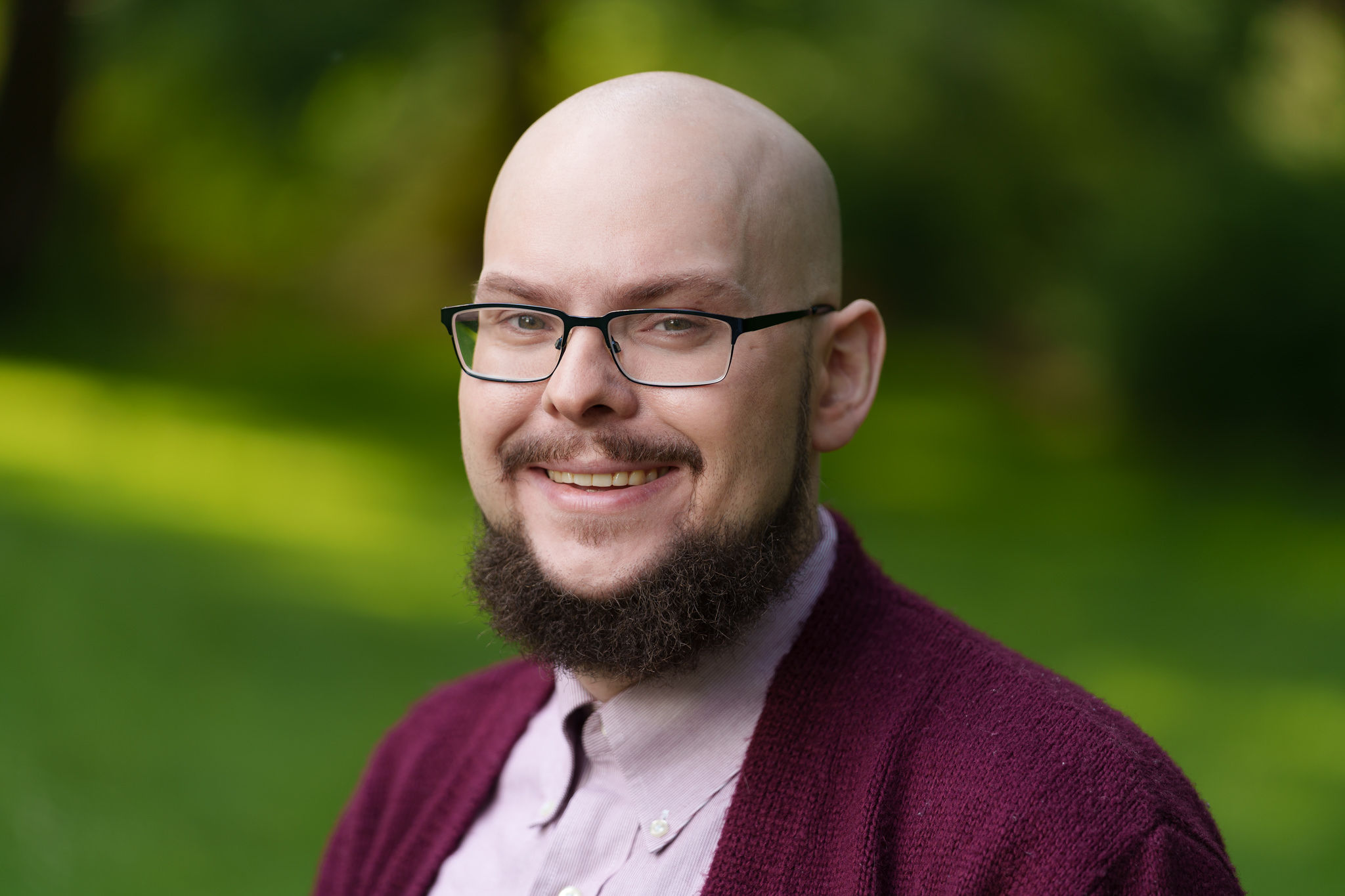 Marshall Short, smiling, wearing glasses and a burgundy cardigan, in front of greenery